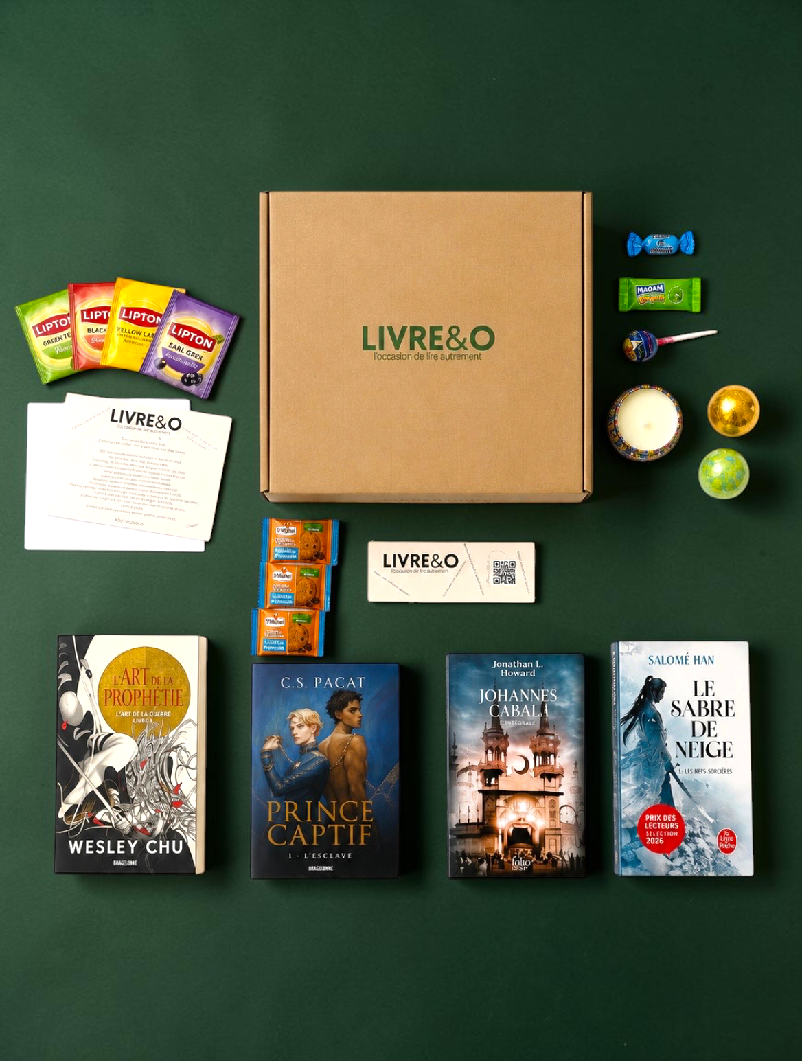 Box Signature Fantasy LIVRE&O contenant quatre romans, des sachets de thé, des friandises et une bougie sur fond vert.