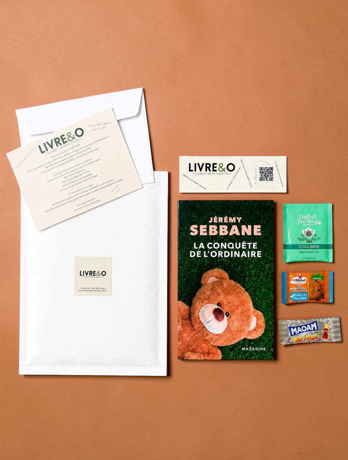 Enveloppe LIVRE&O contenant un roman broché, un marque-page, du thé et des gourmandises sur fond beige.