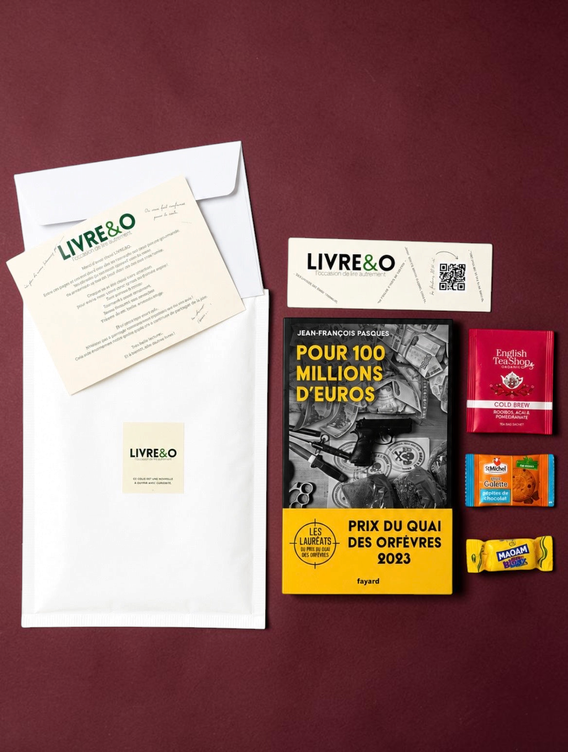 Enveloppe LIVRE&O contenant un roman noir, un marque-page, du thé et des gourmandises sur fond bordeaux